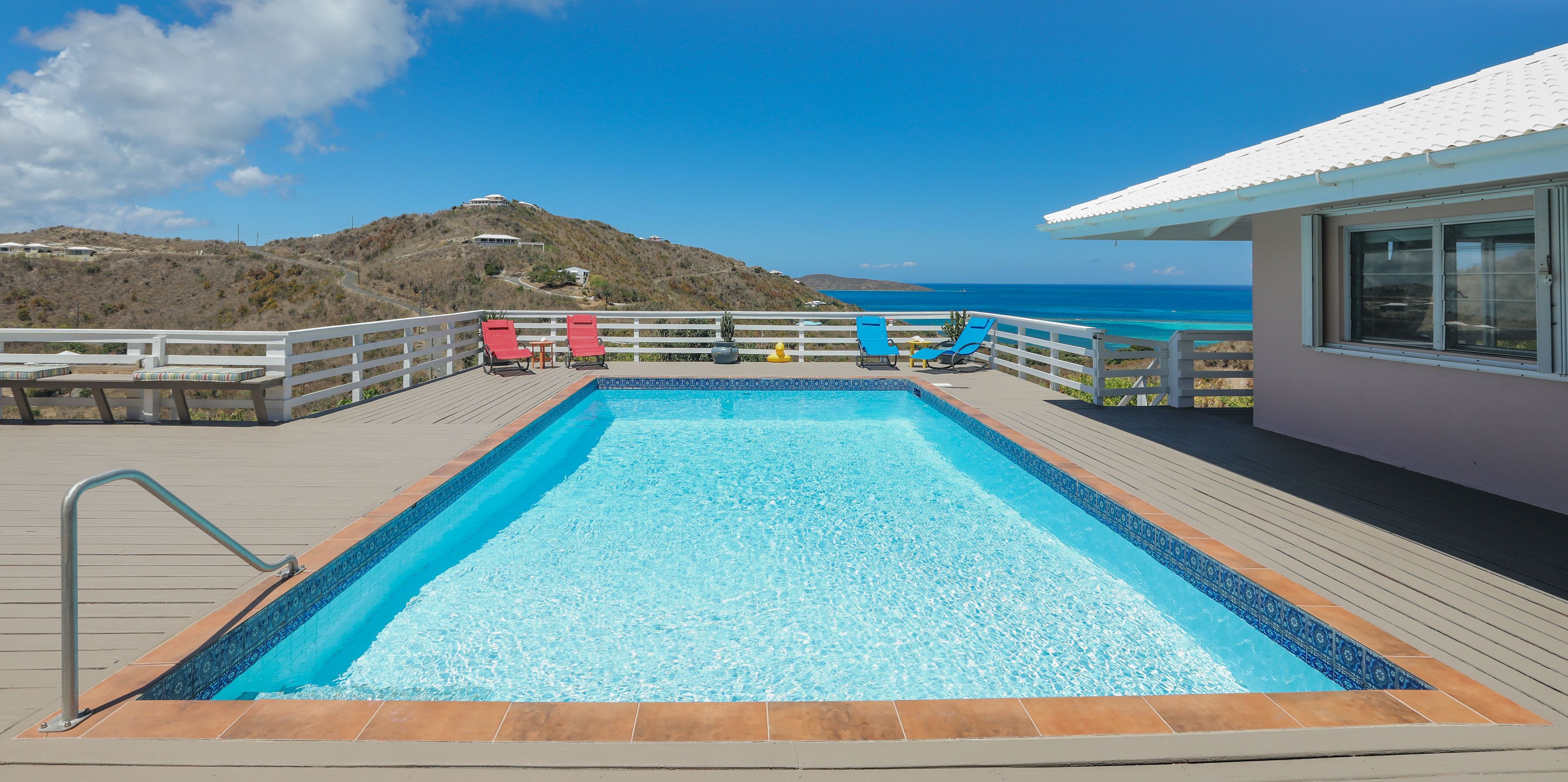 A swimming pool with a deck, lounge chairs, and a view of the ocean and hills.