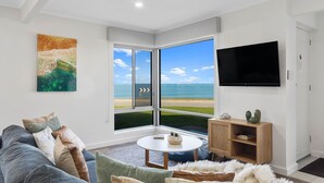A living room with a large window offering a view of the beach, a flat-screen TV mounted on the wall, and a cozy seating area with a sofa and cushions.
