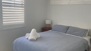 A neatly made bed with a grey bedspread and pillows, a bedside lamp on a wooden nightstand, and a window with white blinds.
