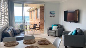 A living room with a sofa, armchairs, a TV, and a table with placemats. There is a balcony with a view of the sea.