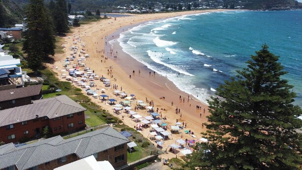 A beach with numerous umbrellas and people, surrounded by residential buildings and trees.