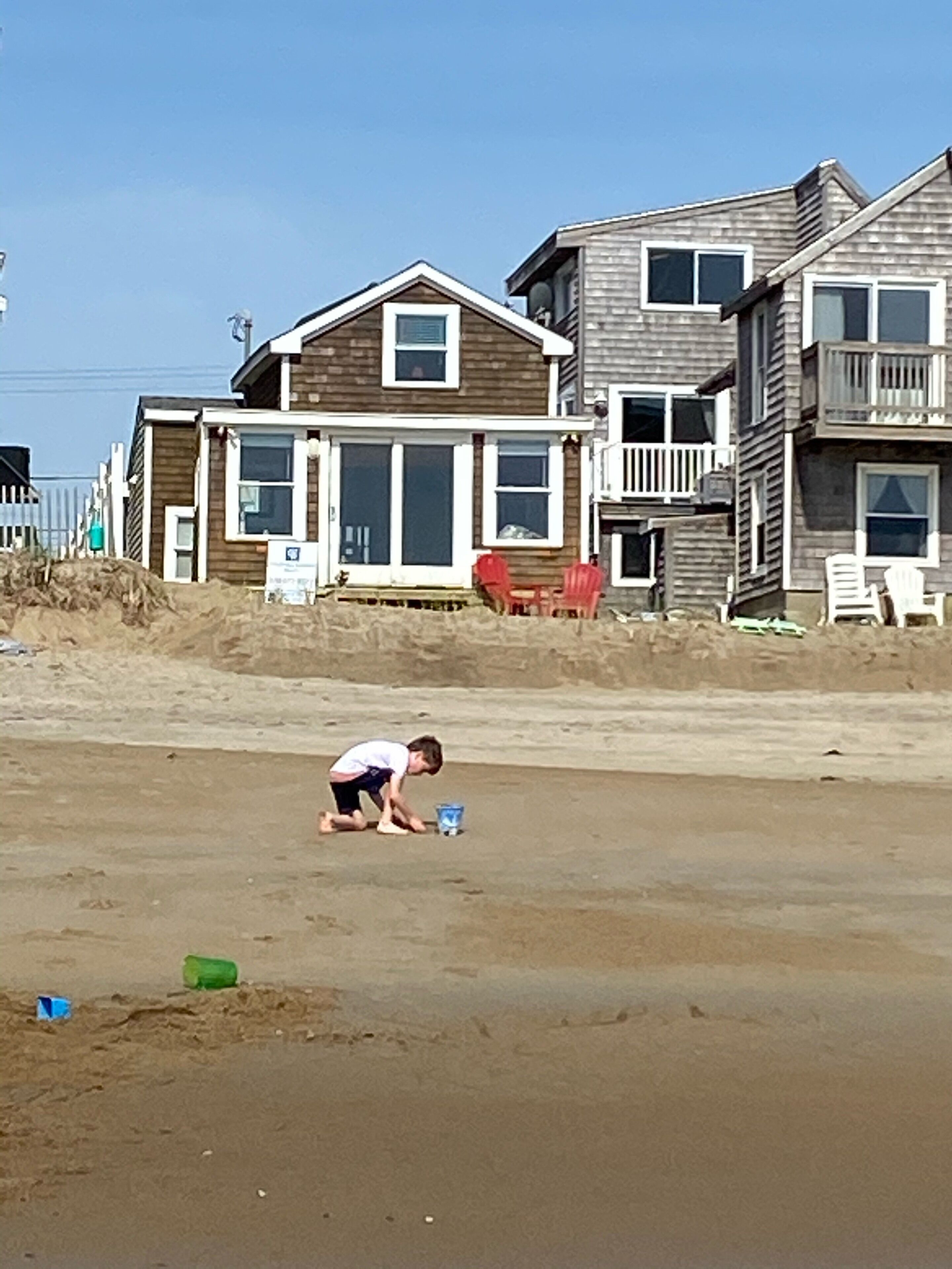 A beachfront property with a sandy beach, a wooden house with a balcony, and a child playing.