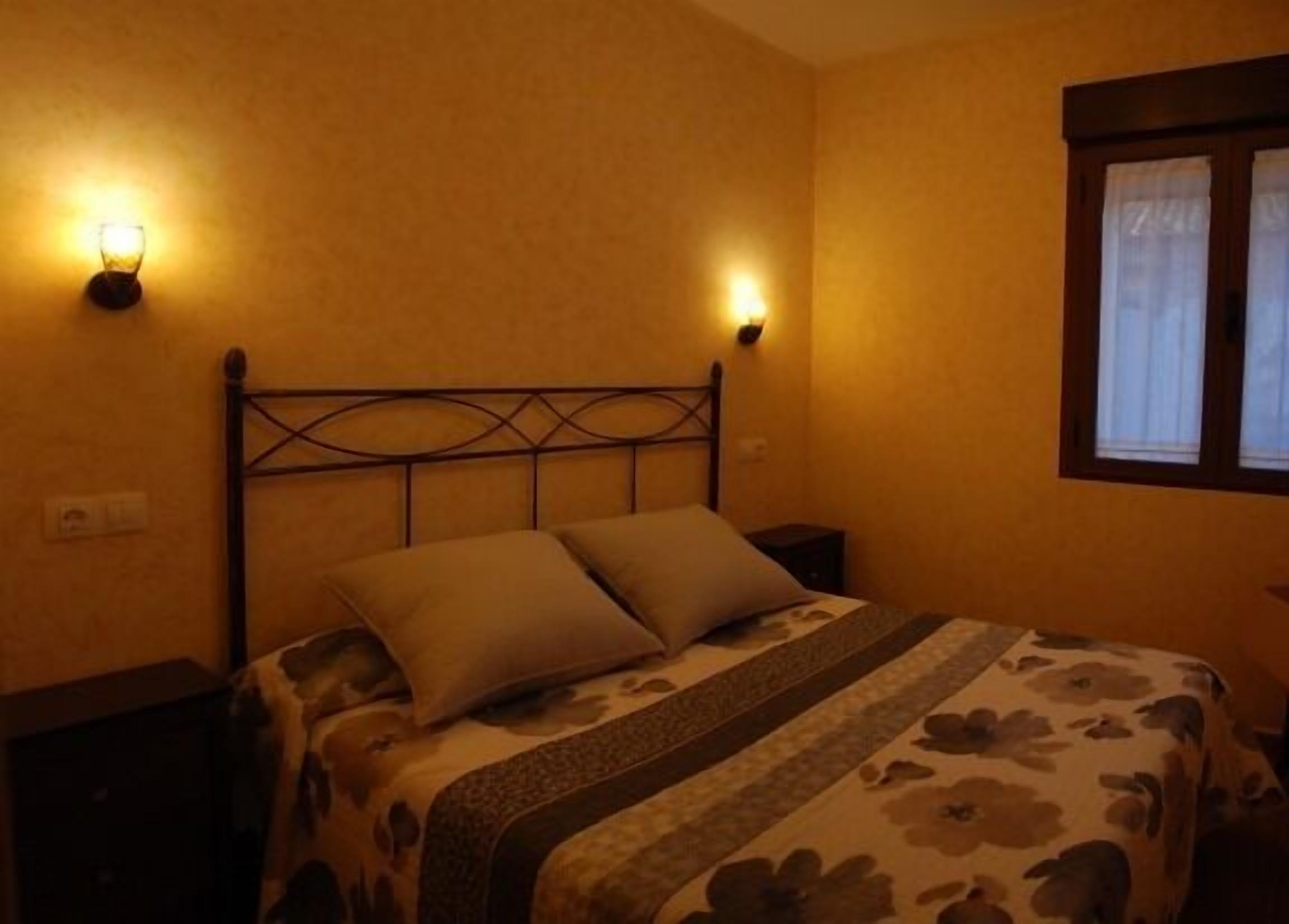 A room with a bed, two wall-mounted lights, a window with curtains, and a nightstand.
