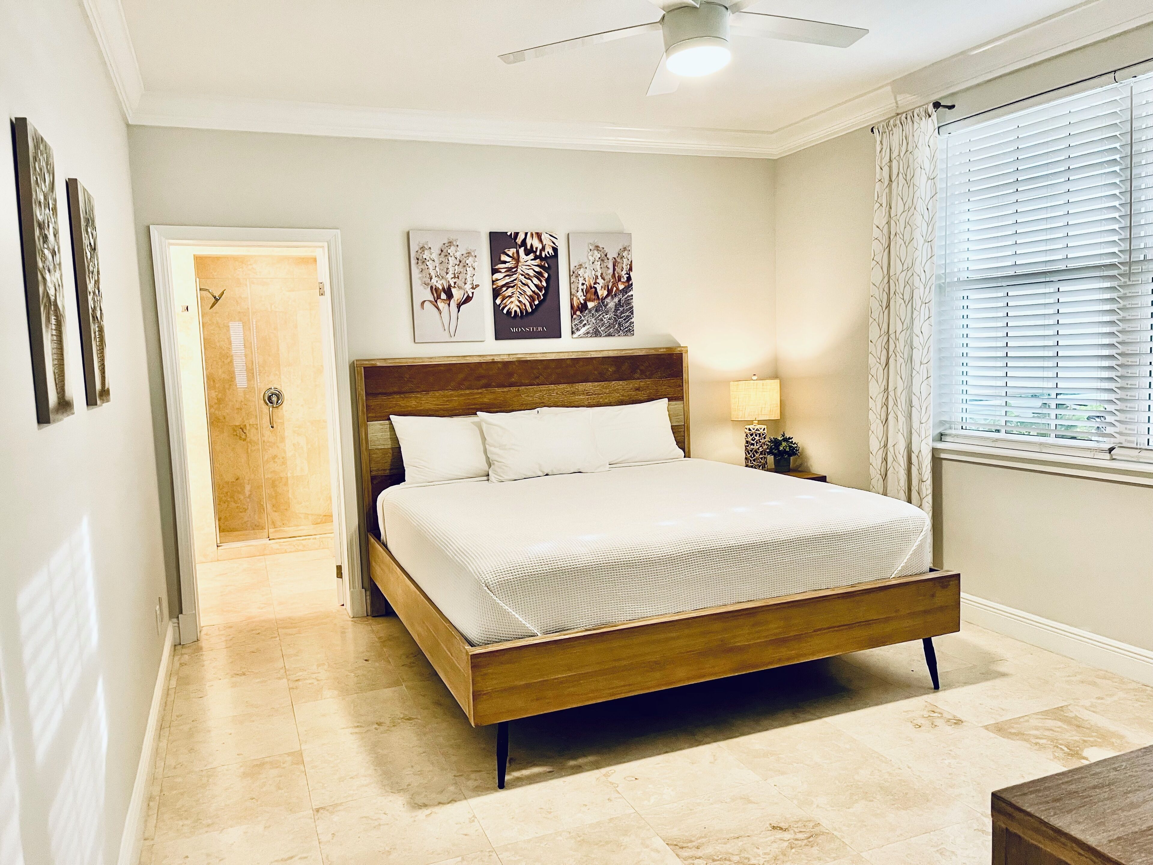 Un dormitorio con cama de madera, ventilador de techo, mesita de noche con lámpara y ventana con persianas.