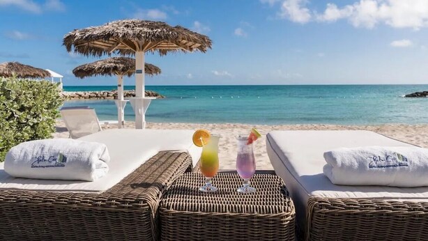 Een strandlounge met witte ligstoelen, twee rieten parasols en twee drankjes op een rieten tafeltje.