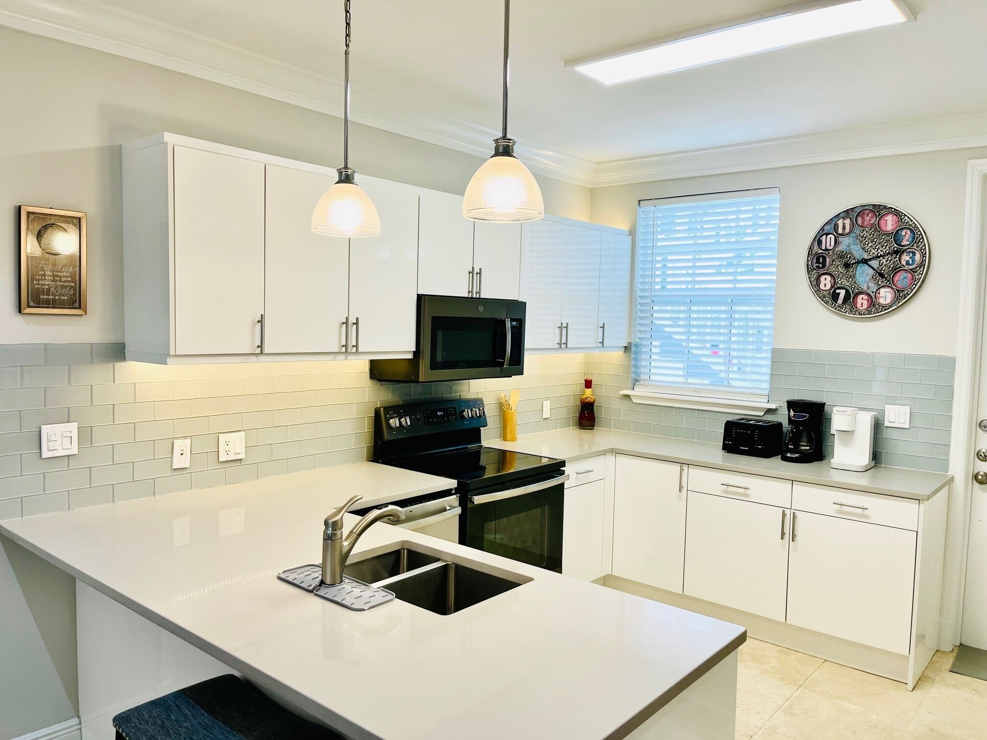 A modern kitchen with white cabinets, a central island with a sink, a microwave, and a stove. There are pendant lights, a window with blinds, and a decorative clock on the wall.