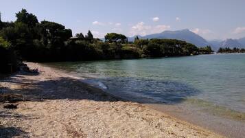 Platja de pedres amb aigua cristal·lina, arbres i muntanyes al fons.