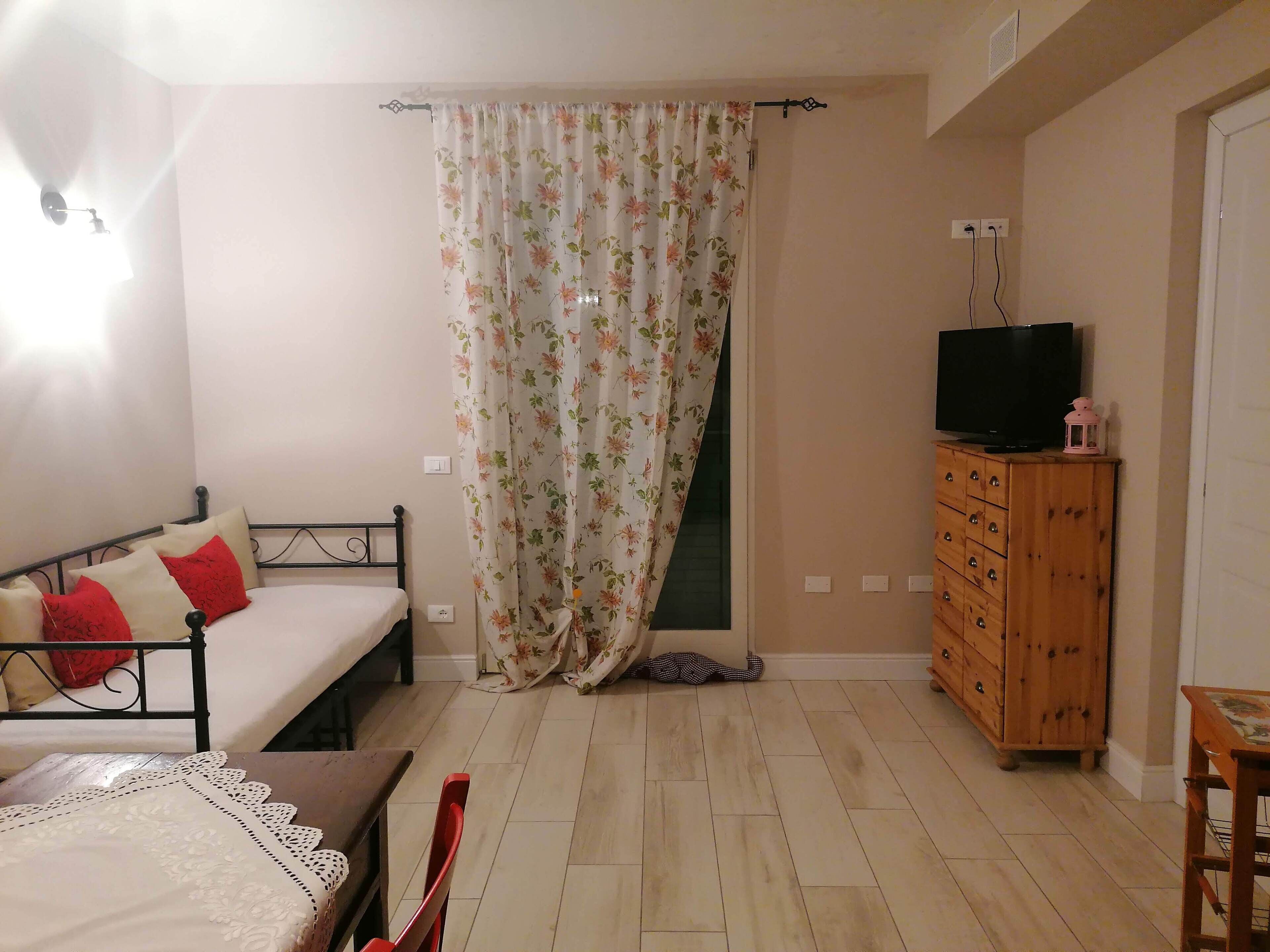 A bedroom with a bed, a wooden dresser, a television, and a window with floral curtains.