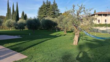 Zona verda ben cuidada amb un hamaca, arbres i un edifici al fons.