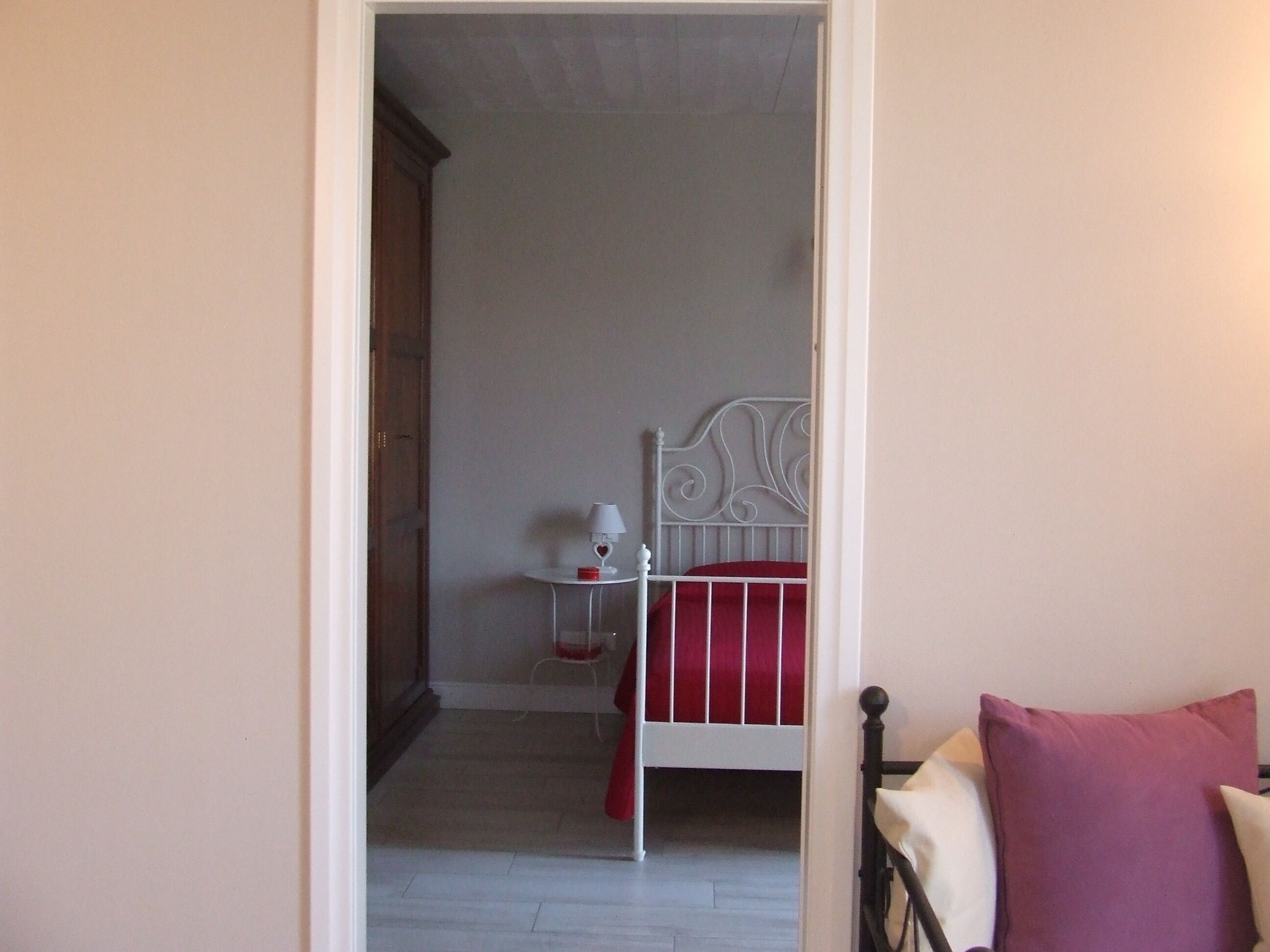 A bedroom with a white metal bed frame, a red and white striped mattress, a small bedside table with a lamp, and a red pillow.