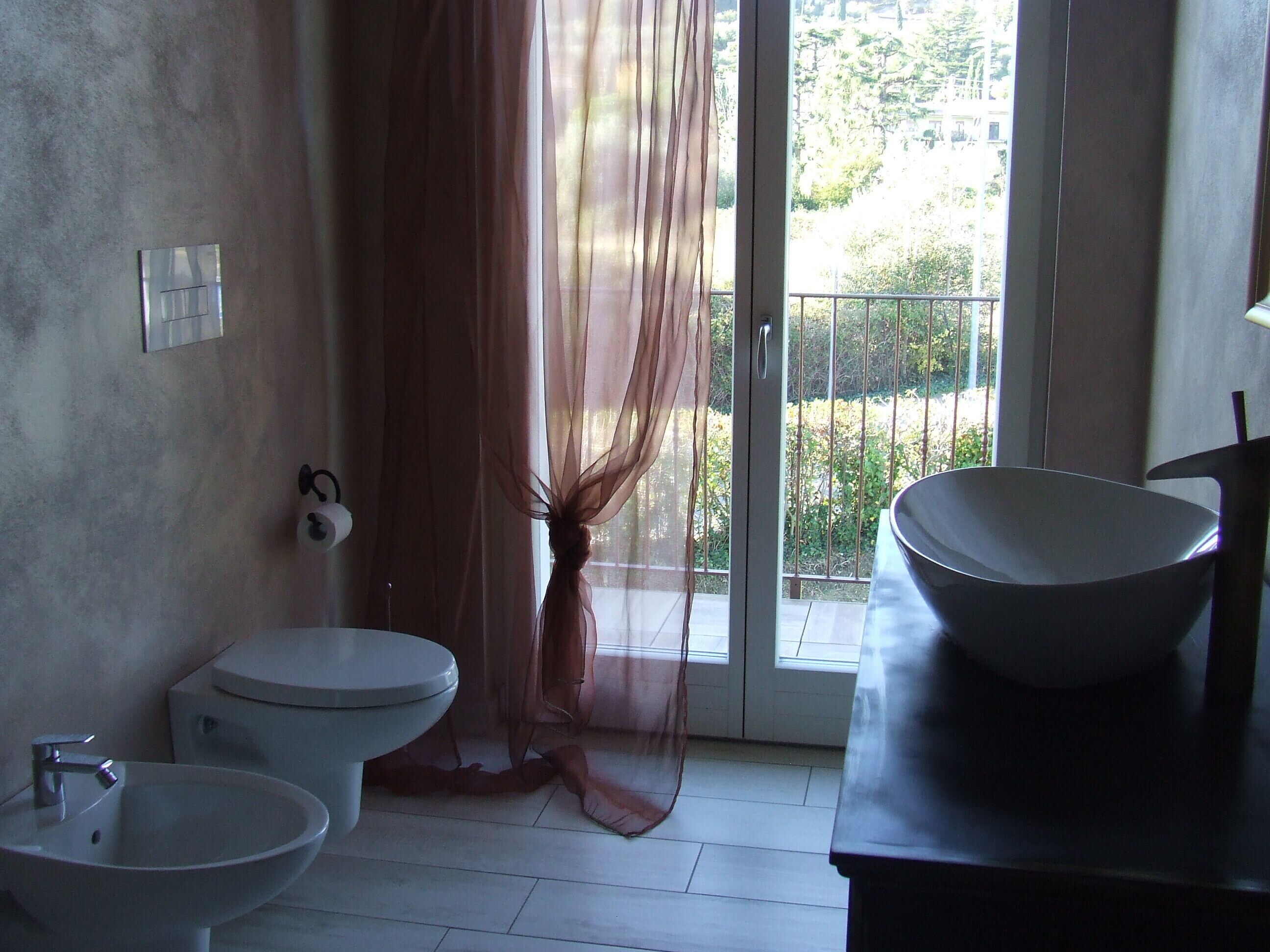 Ein Badezimmer mit Toilette, Waschbecken und einem großzügigen Waschtisch. Ein Vorhang ist teilweise gezogen, und durch das Fenster ist ein Balkon mit Geländer zu sehen.
