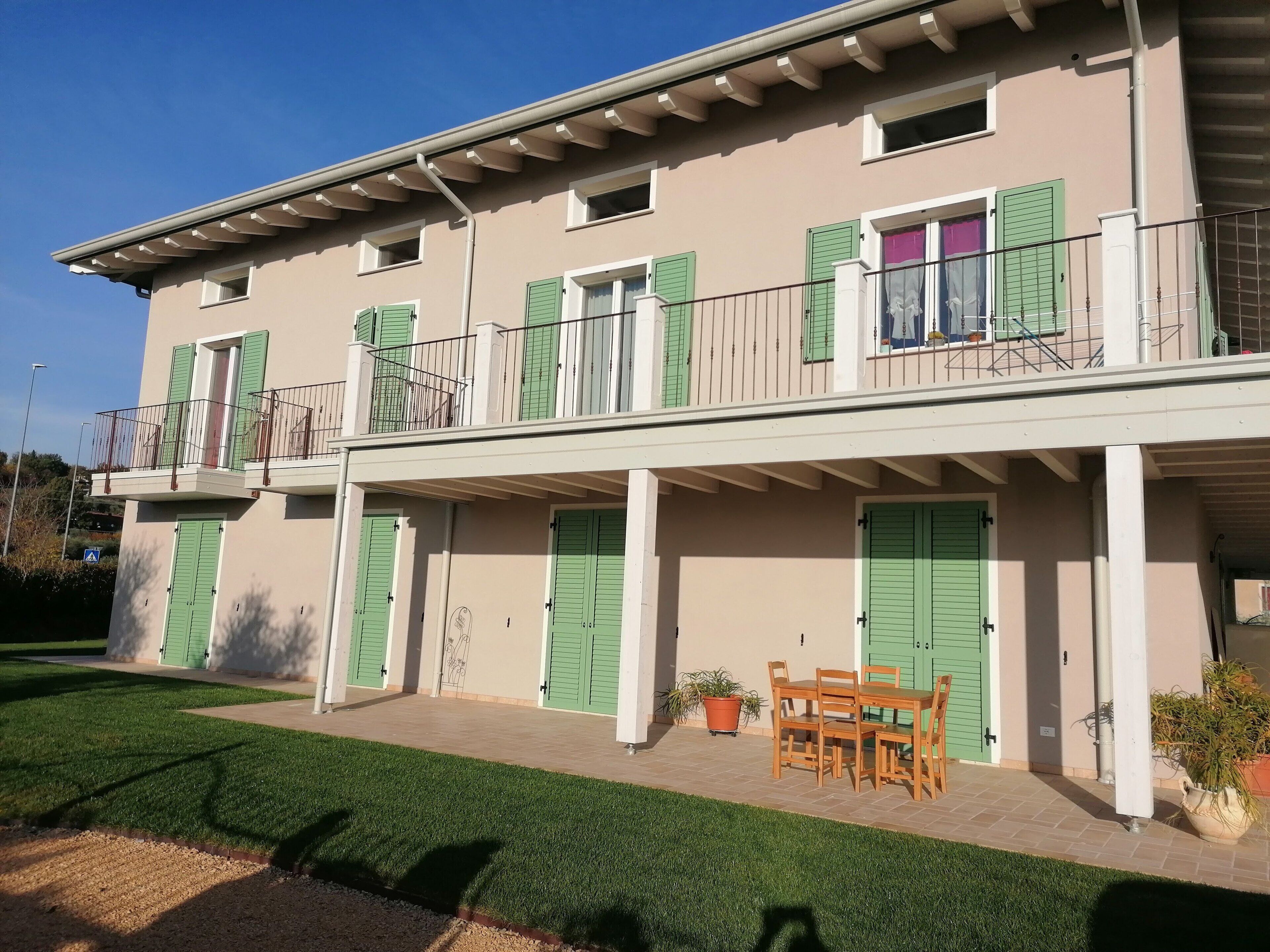 Un bâtiment à deux étages avec des volets verts, un balcon et un petit espace extérieur aménagé pour s’asseoir.