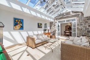 A spacious room with a high ceiling and skylight, featuring a wicker sofa, a glass coffee table, and a dining area with a stone wall. - Coedrath House - Luxury Manor, Swim Spa, Sea Views (Saundersfoot)
