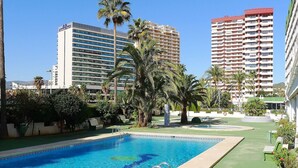 A swimming pool with a clear blue water, surrounded by palm trees and greenery, with high-rise buildings in the background. - Beautiful apartment for 4 people with A/C, pool, WIFI, TV and terrace (Calpe)