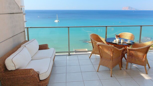 Ein Balkon mit einer weißen Couch und Korbstühlen mit Blick auf das klare, blaue Meer und ein Segelboot.