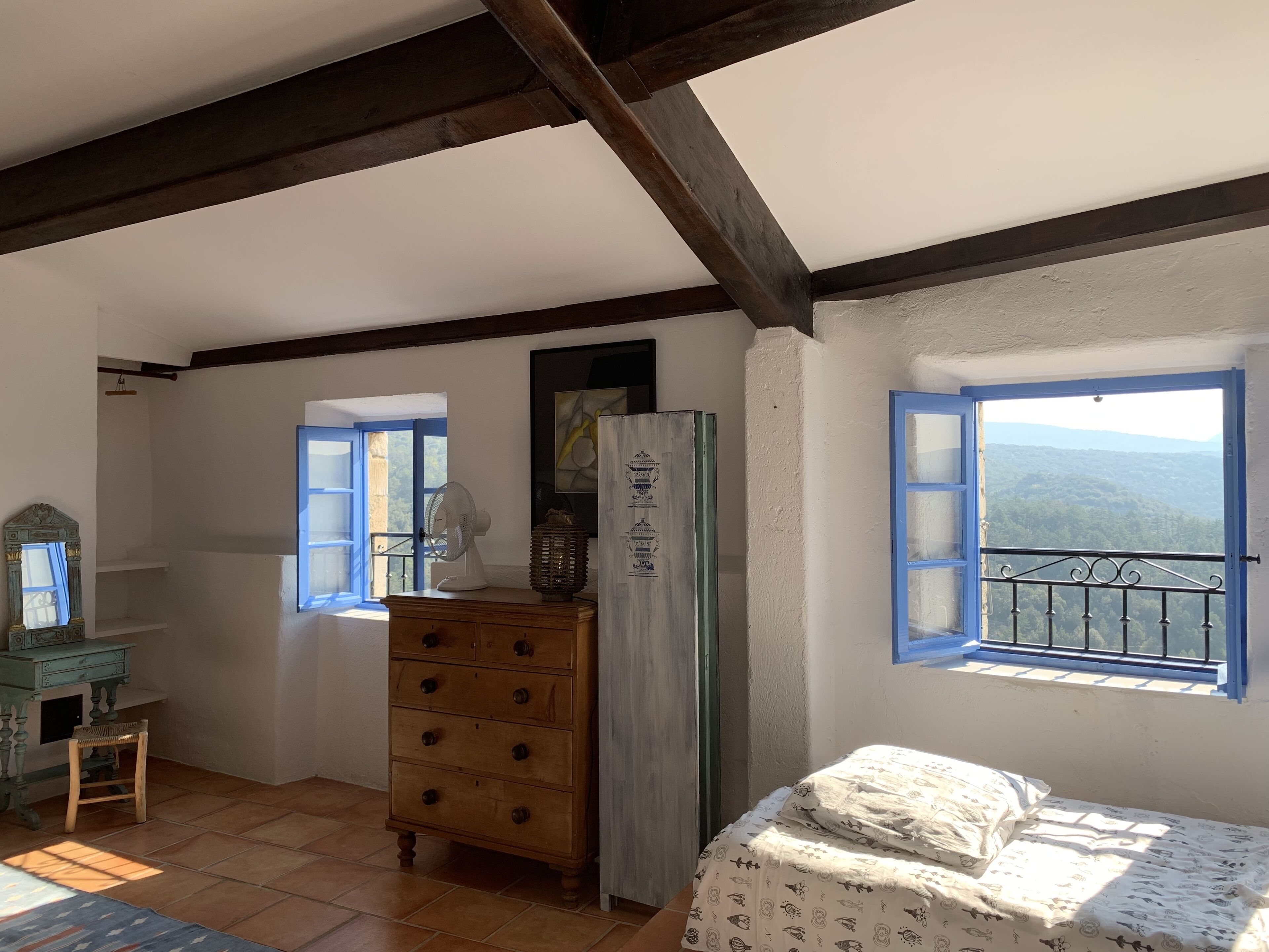 A room with a bed, a dresser, a fan, and a window with a view of the mountains.
