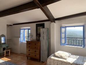 Une chambre avec un lit, une commode, un ventilateur et une fenêtre donnant sur les montagnes.