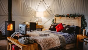 Ein gemütliches Zelt mit Kaminofen, einem Bett mit rotem Kopfkissen, einem Schreibtisch mit Lampe und einem kleinen Tisch mit Decke.