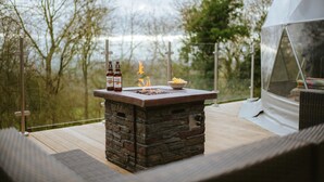 Ein Außenbereich mit Bar, zwei Bierflaschen, einer Schüssel mit Snacks und einem Lagerfeuer.