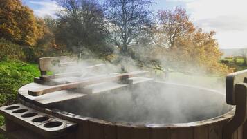 Un jacuzzi con estructura de madera y un banco, rodeado de árboles y una zona verde.