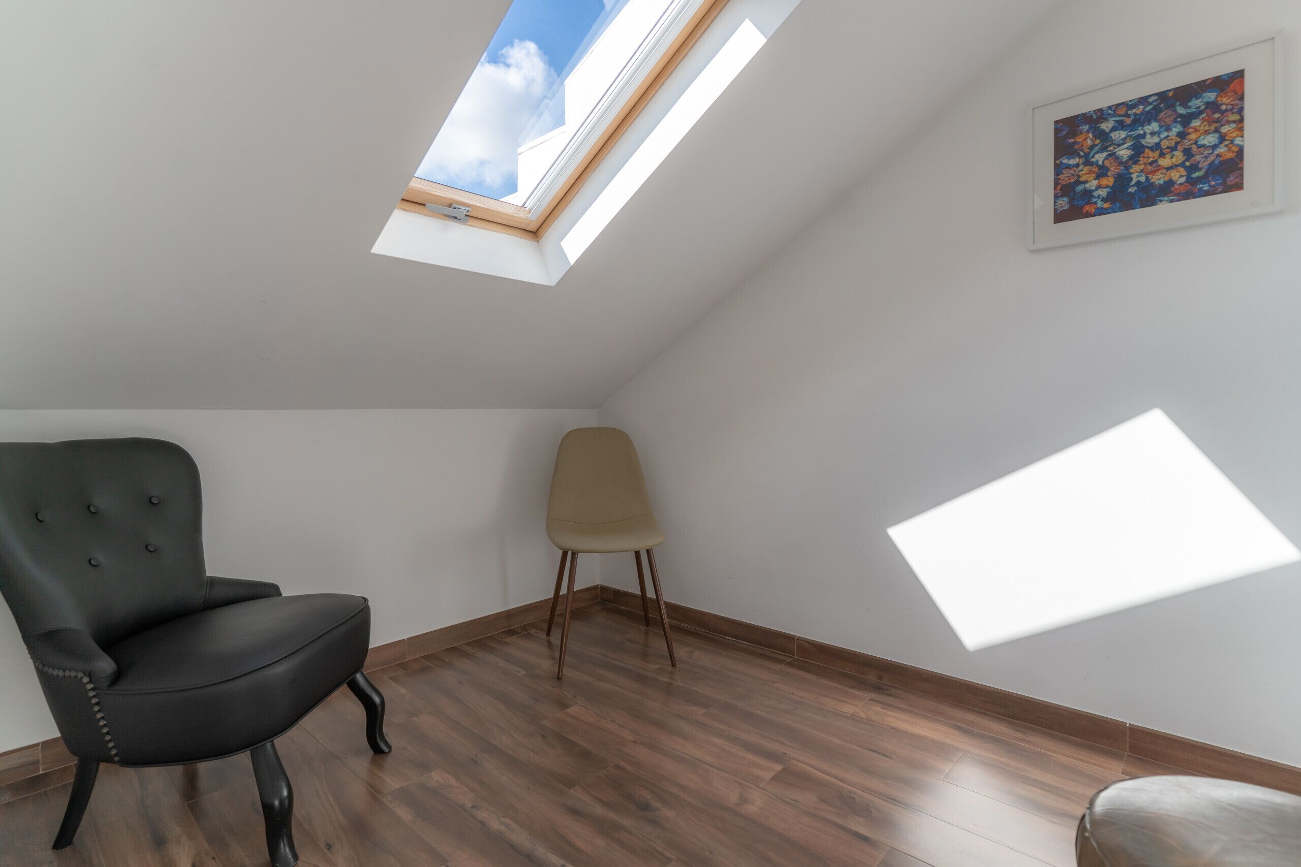 Une pièce avec un plafond en pente, un puits de lumière, une chaise noire, une chaise beige et un plancher en bois.