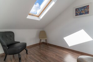 Una habitación con techo inclinado, tragaluz, una silla negra, una silla color beige y piso de madera.