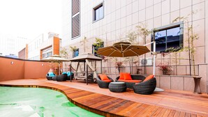 Un área de piscina en la azotea con cubierta de madera, asientos al aire libre y un gran paraguas.