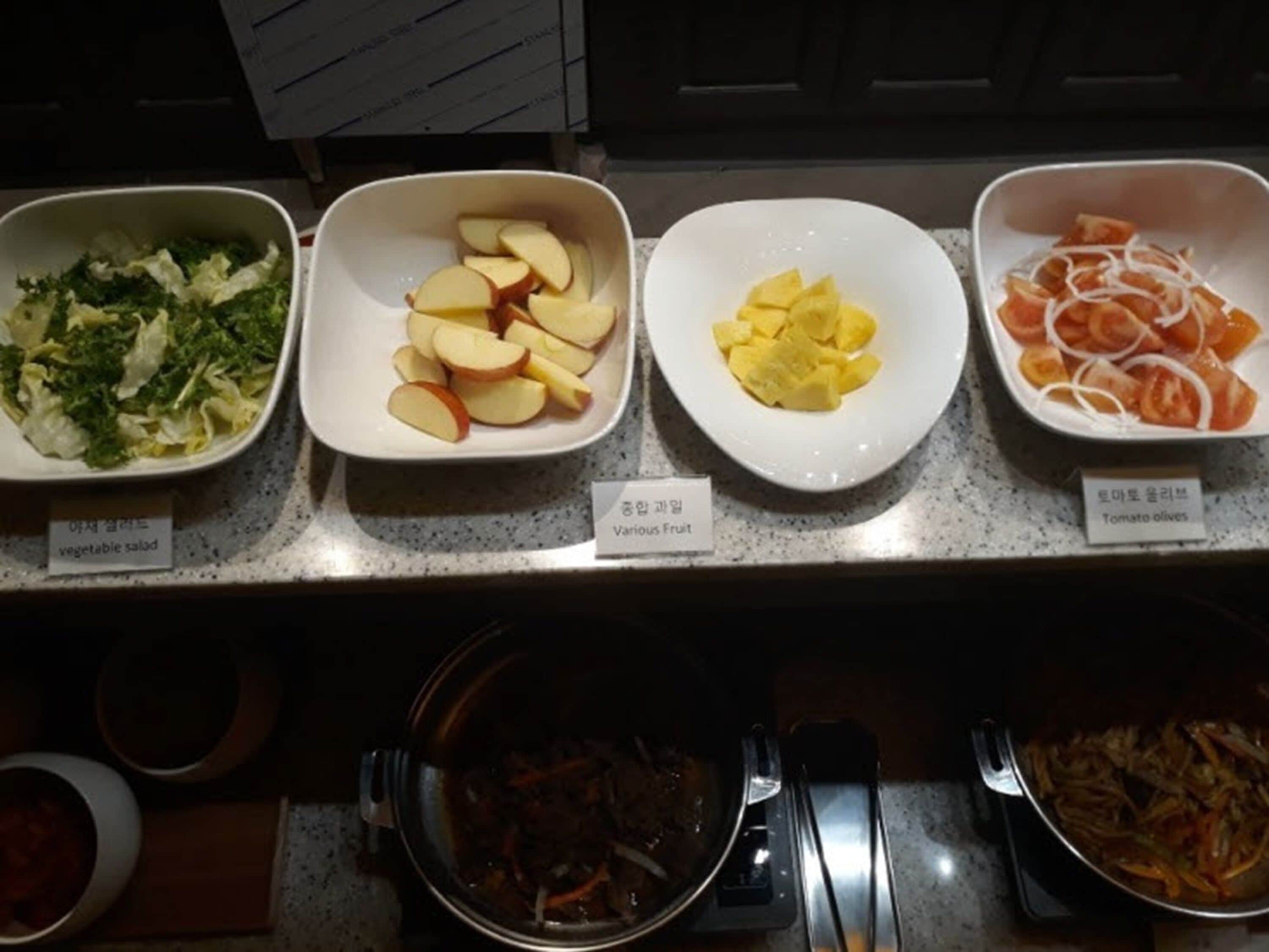 Un bufé con diversos platos, incluyendo ensalada de verduras, manzanas, uvas y aceitunas de tomate.