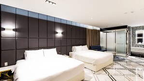 Habitación de hotel con dos camas, un cabecero grande y oscuro, un vestidor amplio con paredes de vidrio, y un baño moderno con piso de baldosas blanco y negro.