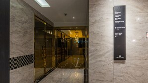 Un vestíbulo de hotel con piso de mármol, puertas de vidrio que dan a un área de descanso y un letrero que enumera servicios como desayuno, cafetería, sala de descanso, sala de negocios, lavandería y habitaciones de huéspedes 201 a 206.