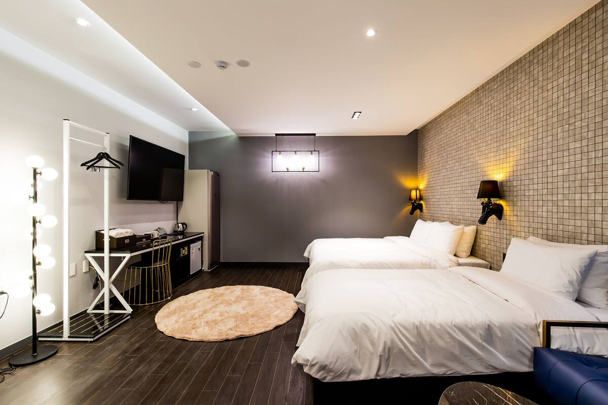 Ein modernes Hotelzimmer mit zwei Betten, einem Schminktisch mit Beleuchtung, einem Flachbildfernseher und einer Wandleuchte.