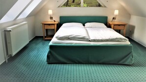 Ein Schlafzimmer mit schräg steilender Decke, einem Bett mit weißen Bettwäsche, zwei Nachttischen mit Lampen und einem Fenster mit Blick auf Bäume.