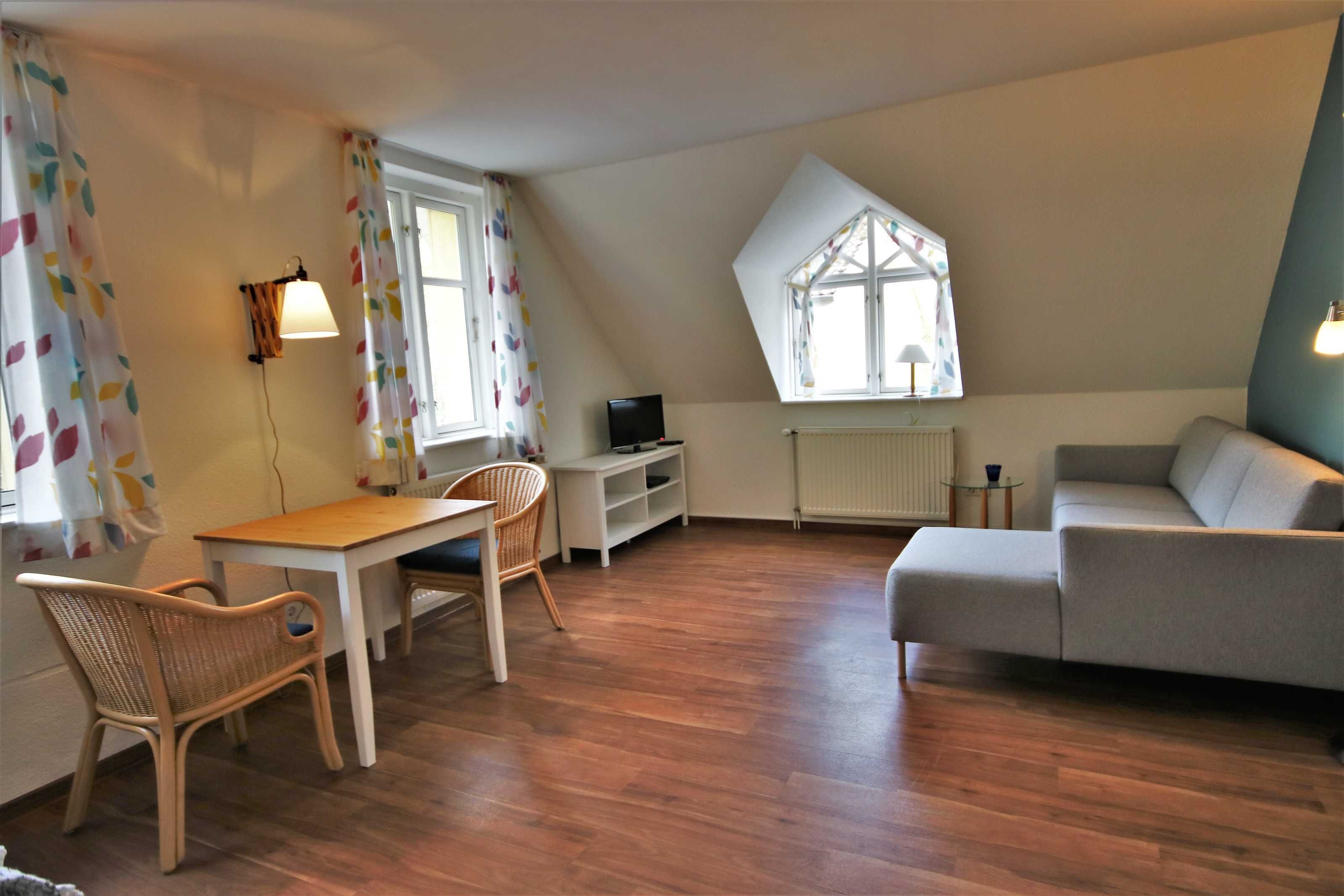 A room with a wooden floor, a sofa, a table, a chair, a TV, and a window with a view.
