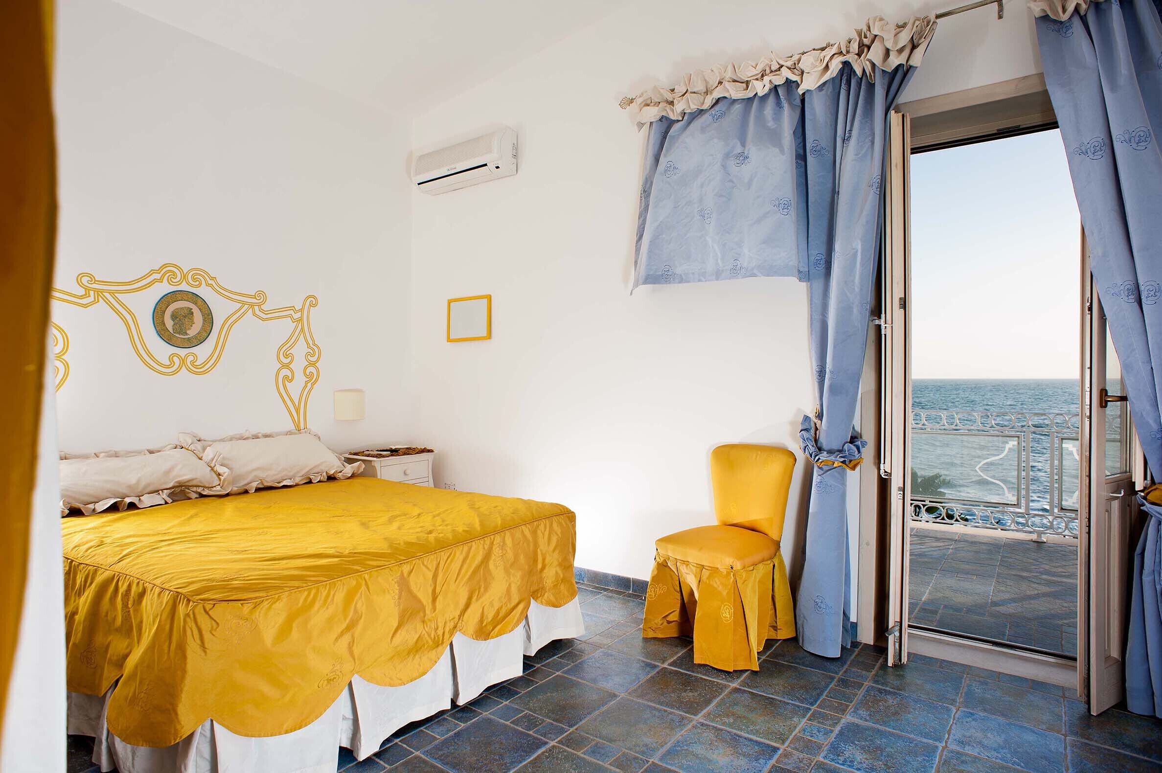 A bedroom with a bed, a chair, a window with curtains, and a view of the sea.