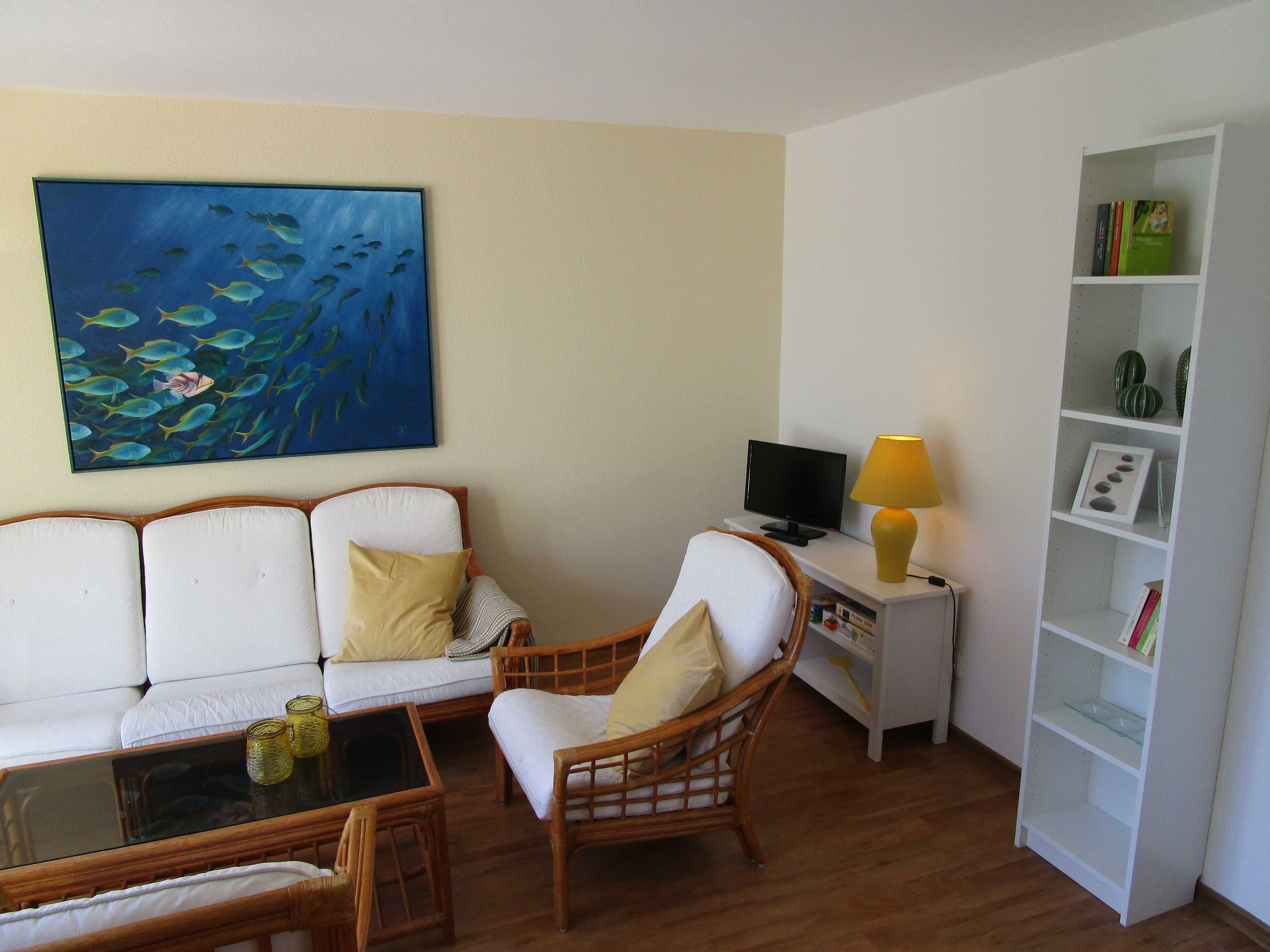 A living room with a white sofa, a wicker chair, a glass coffee table, a TV on a white stand, and a bookshelf.