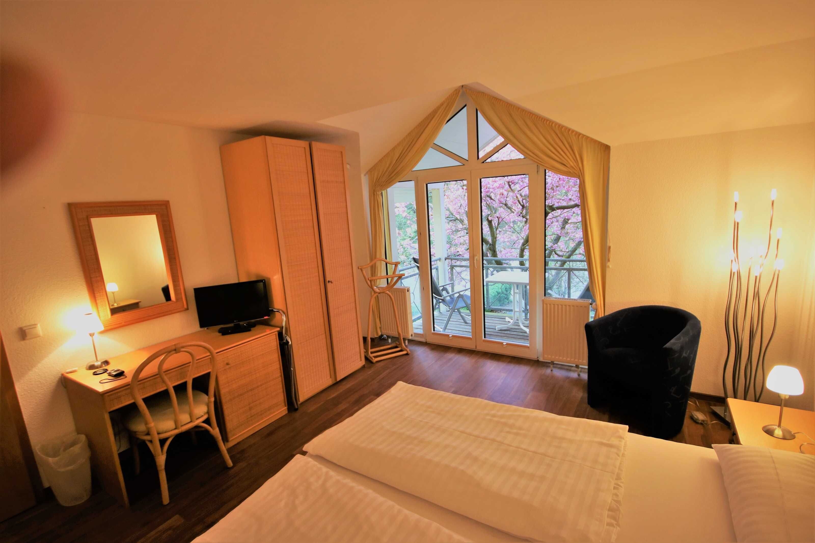 A hotel room with a bed, a desk with a chair, a television, a wardrobe, and a large window with a view of cherry blossoms.
