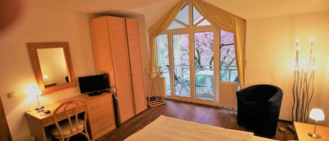A hotel room with a bed, a desk with a chair, a television, a wardrobe, and a large window with a view of cherry blossoms.