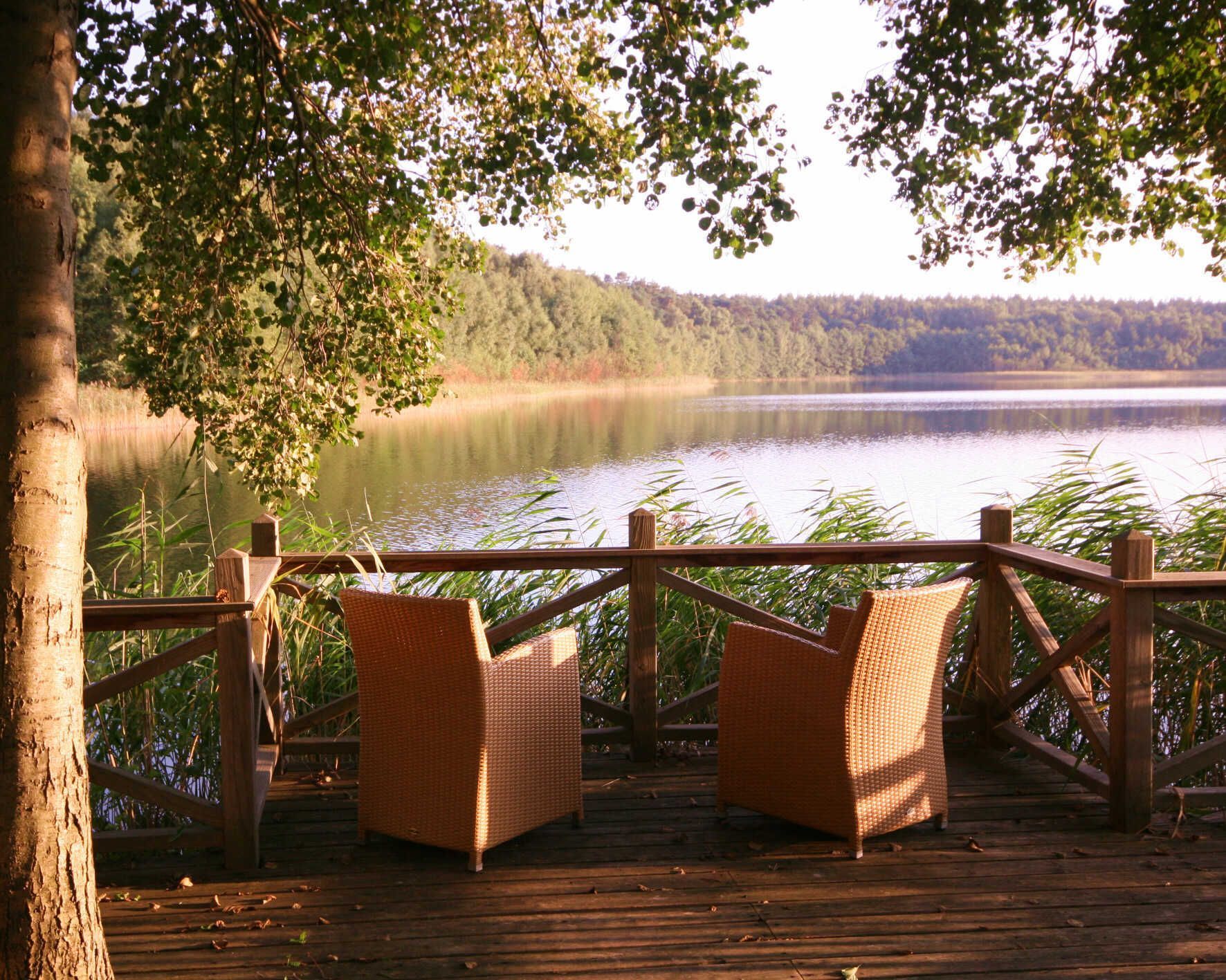 A wooden deck with wicker chairs overlooking a calm lake, surrounded by trees.