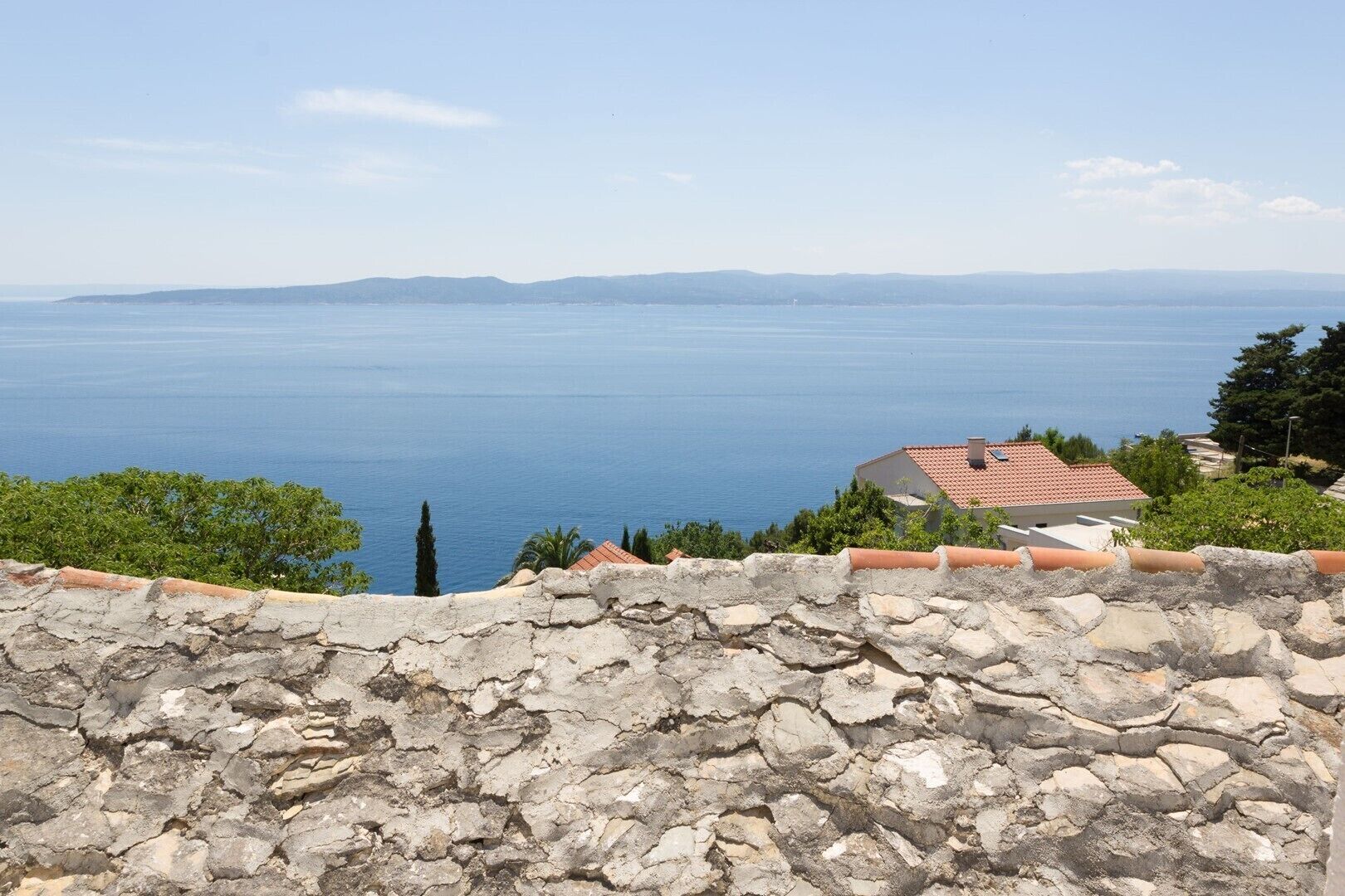 A coastal view with a stone wall in the foreground, a building with a red-tiled roof, and a clear blue sea extending to the horizon.