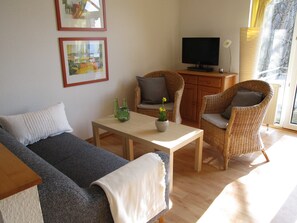 Un salon comprenant un canapé, deux fauteuils en osier, une petite table avec un vase, un téléviseur posé sur un meuble et des tableaux encadrés accrochés au mur.