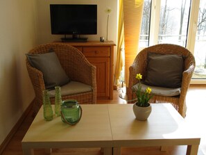 A room with two wicker chairs, a table, a television on a stand, and a window with curtains.