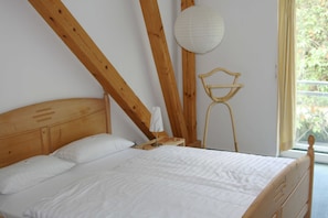 Et soveværelse med sengegavl i træ, hviddynebetræk, et natbord med lampe og et vindue med gardiner.