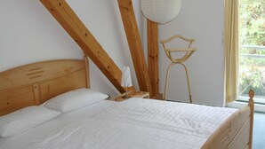 Ein Schlafzimmer mit hölzernem Kopfteil, weißer Bettdecke, einem Nachttisch mit Lampe und einem Fenster mit Vorhängen.