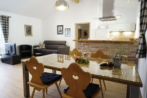 A modern living room with a glass dining table, wooden chairs, a sofa, and a kitchen area with a brick wall.