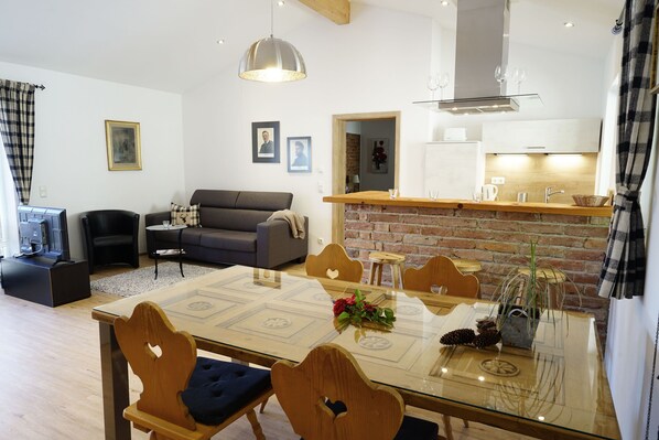 A modern living room with a sofa, a dining table with chairs, a kitchen area with a brick wall, and a hanging light fixture.