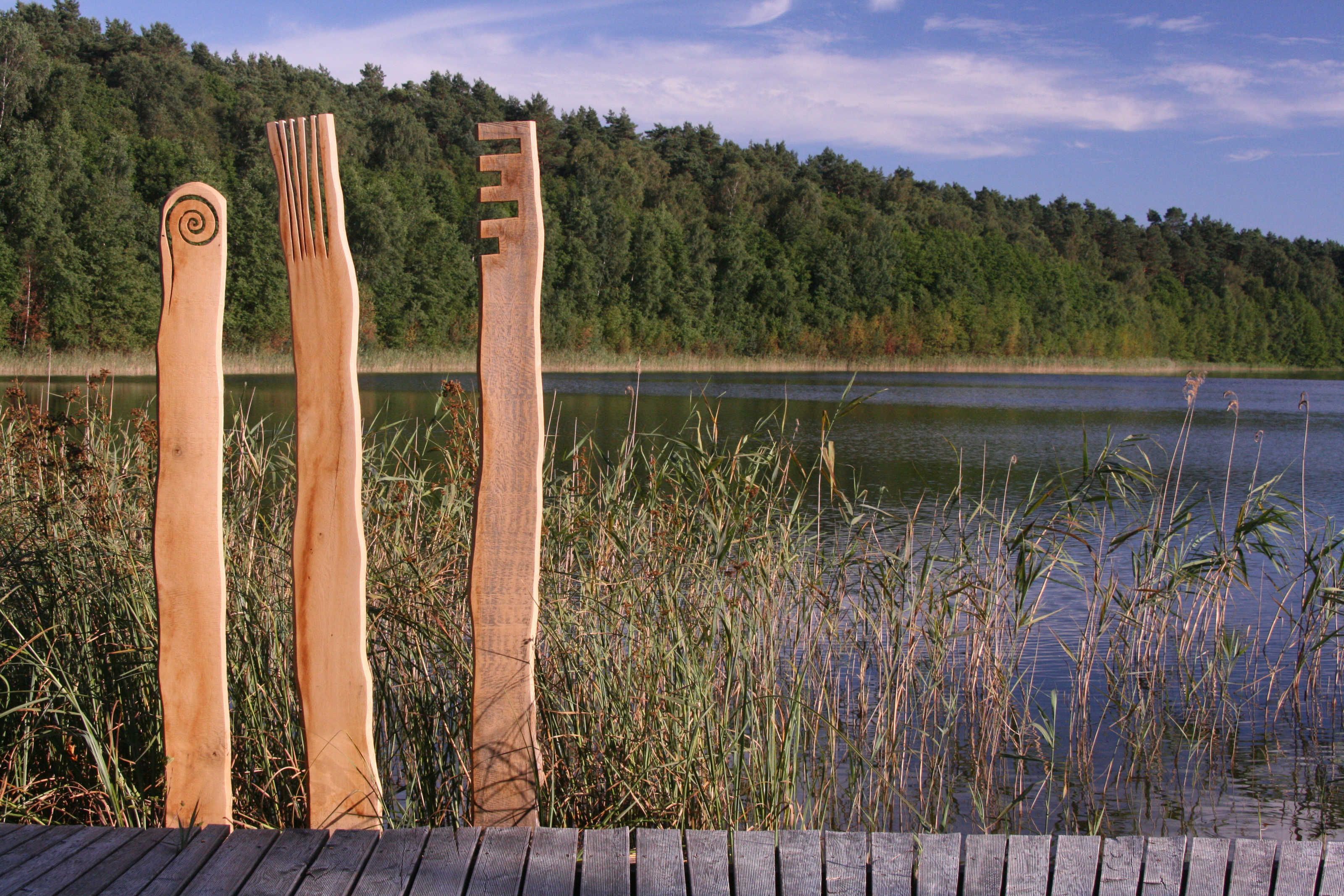 Three wooden sculptures by a lake with a forest in the background.