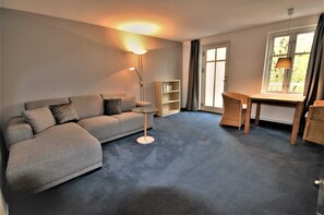 A living room with a sofa, a small table, a bookshelf, and a window with curtains. - KIWI Apartment 8 - KIWI Apartments on Lake Dreier (Alt Schwerin)