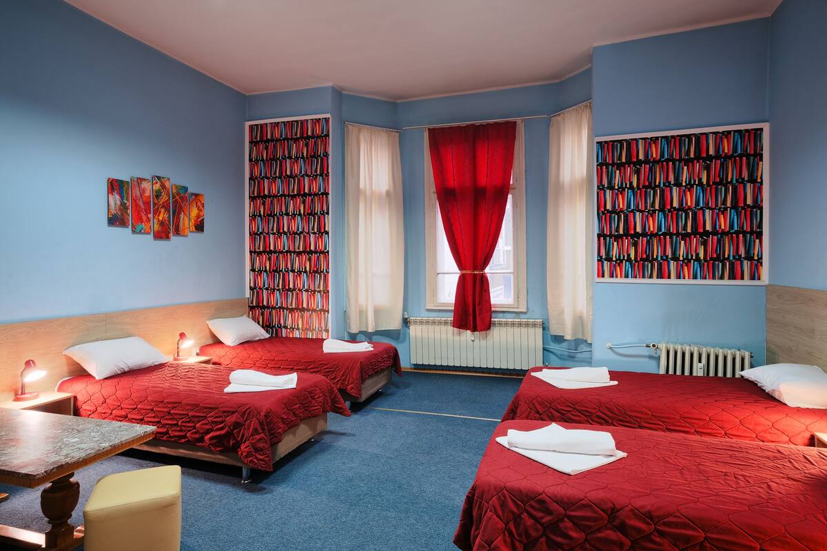 A hotel room with two beds, a bookshelf, a radiator, and a window with red curtains.