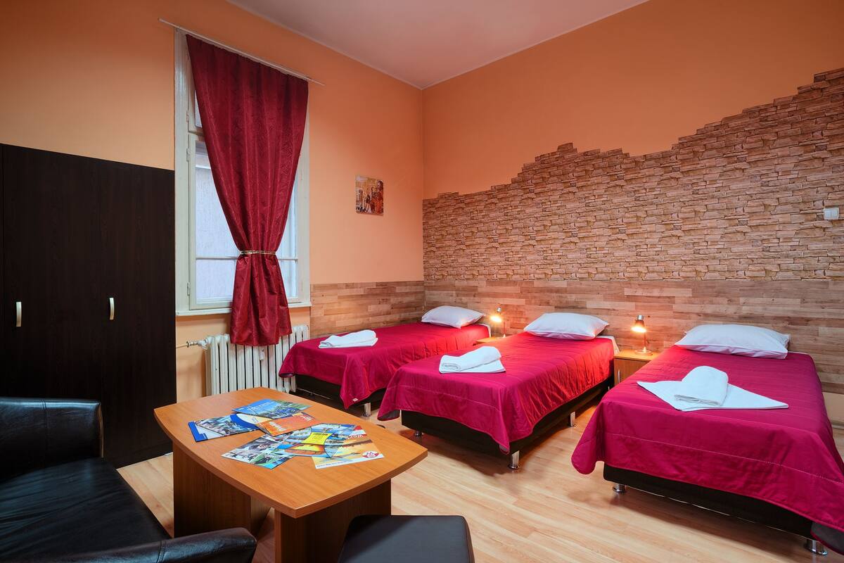 A hotel room with two beds, a wooden table with brochures, a red curtain, and a stone wall.