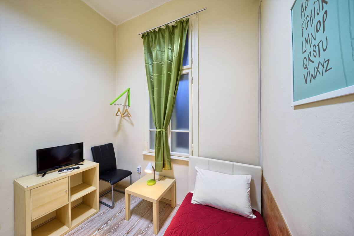 A small, well-lit room with a bed, a desk with a chair, a television, and a window with a green curtain.